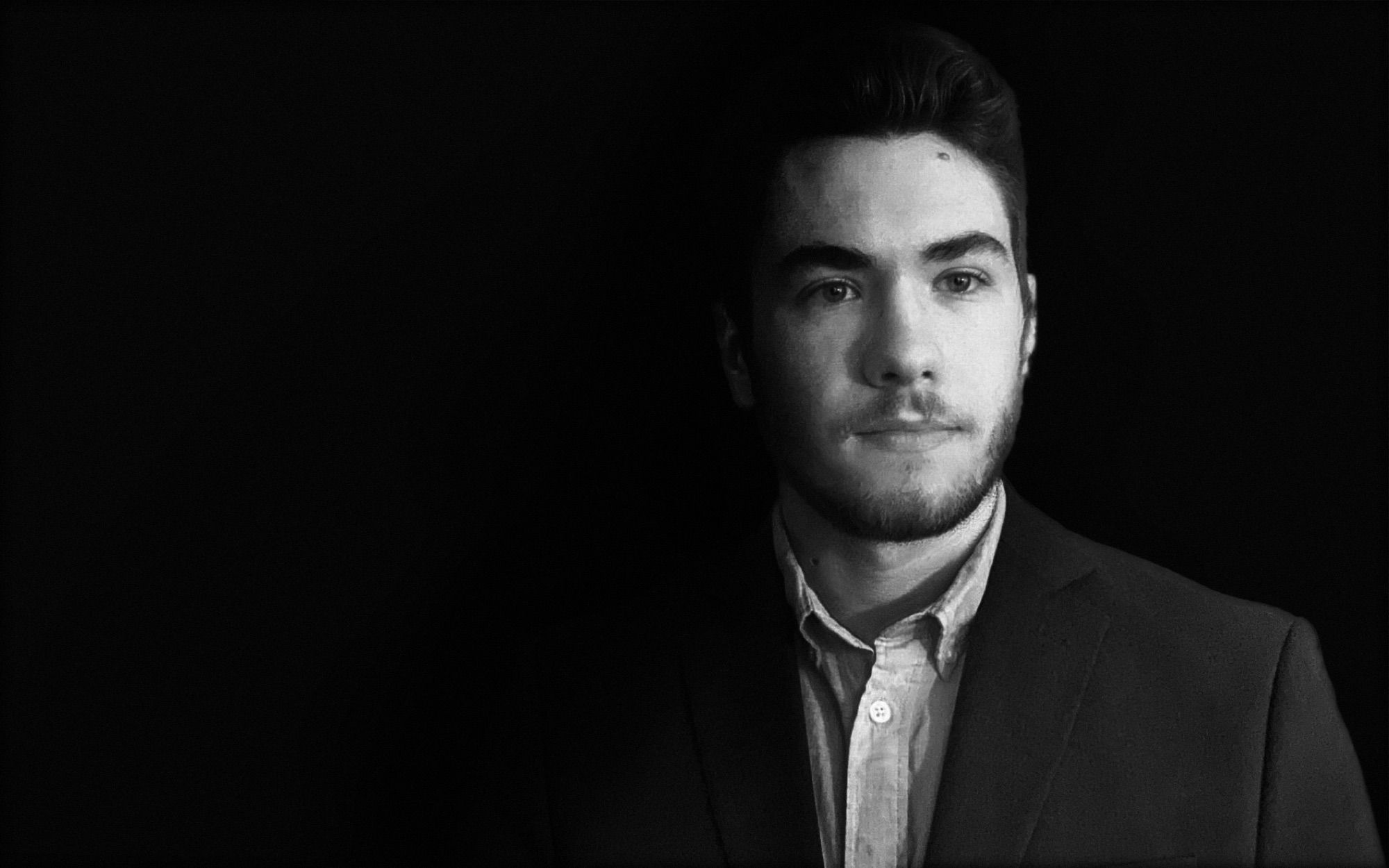 Black-and-white portrait of John Fissel in a dark blazer, lit from the side against a black background